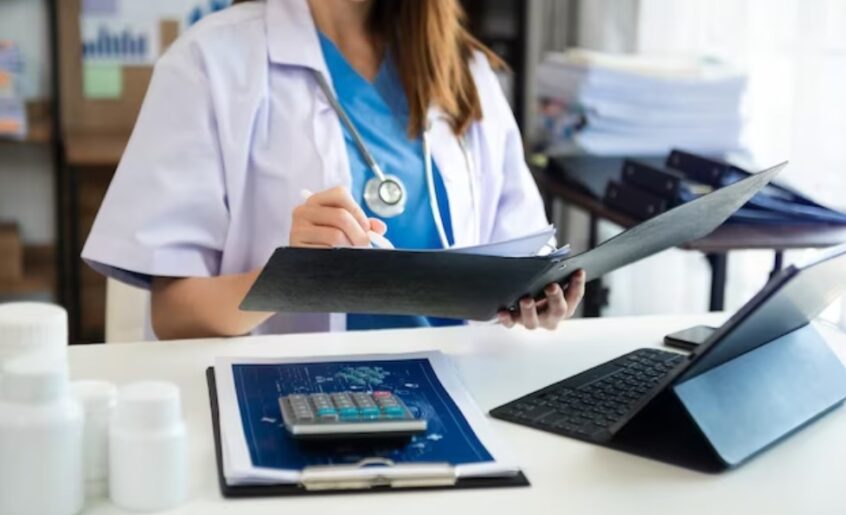 a female doctor working with patient records while sitting at the desk, with a calculator, smartphone, and laptop on it