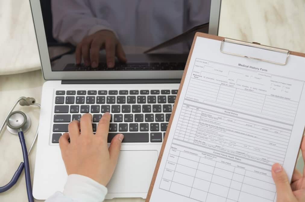 hand typing on the laptop keyboard and holding a sheet with medical information form on it