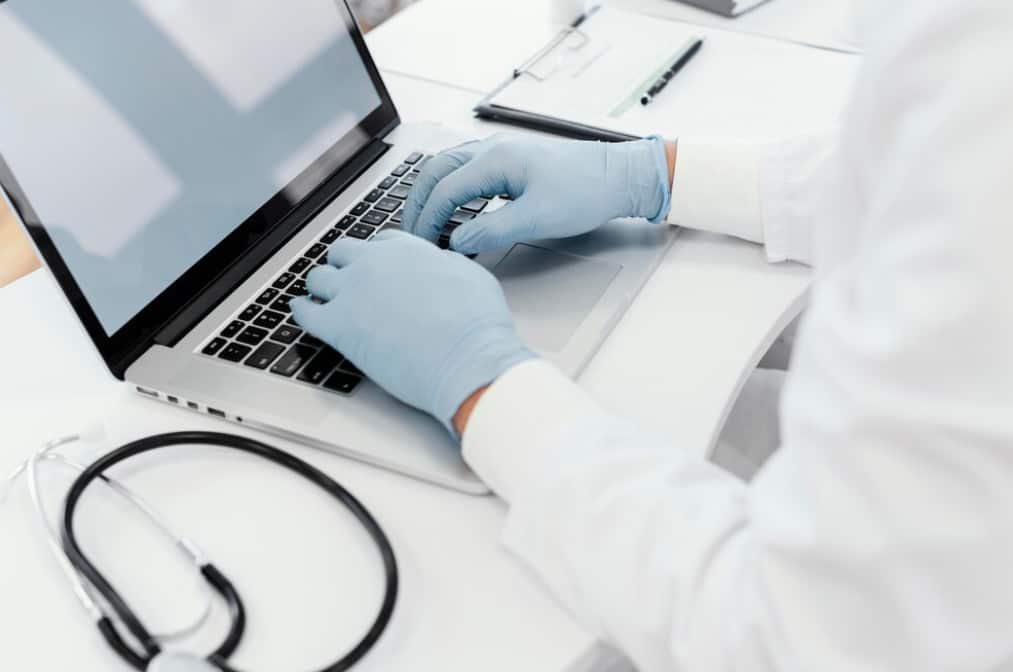 A medic in gloves types on a laptop next to a stethoscope and clipboard