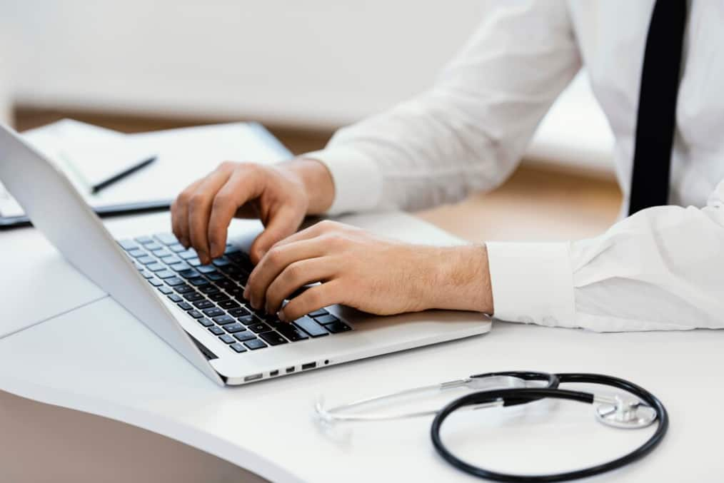 A close-up of a doctor's hands typing on a laptop keyboard, stethoscope nearby