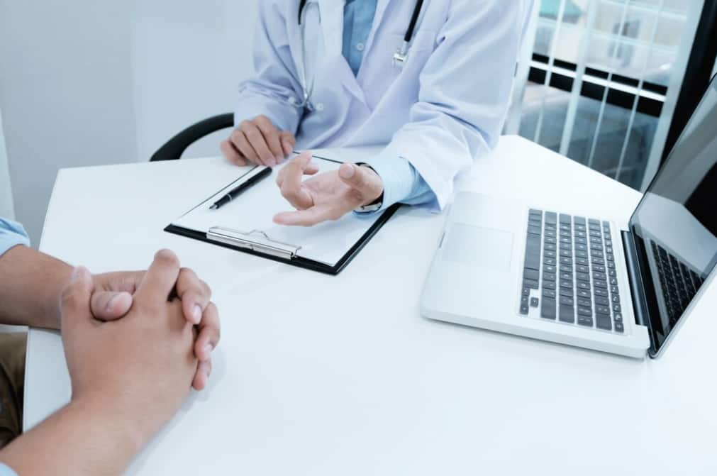 A doctor is discussing with a patient across a desk with a laptop open