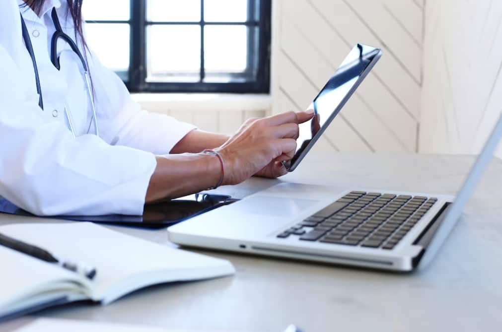 A doctor using a tablet next to a laptop on a sunny desk