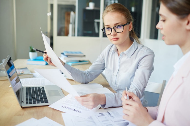 two young women sitting at a desk in an office with documents in their hands